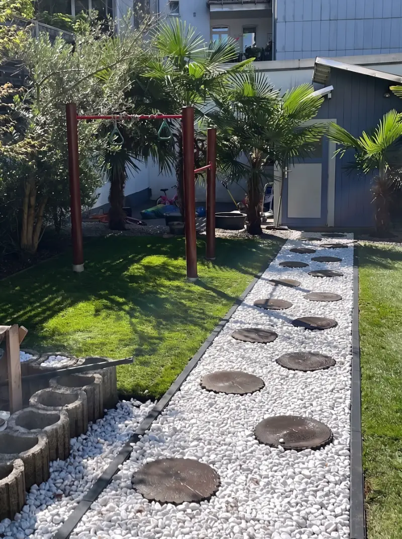 Sonniger Garten mit Rasen, Palmen und einem Weg aus weißem Kies mit runden Trittplatten