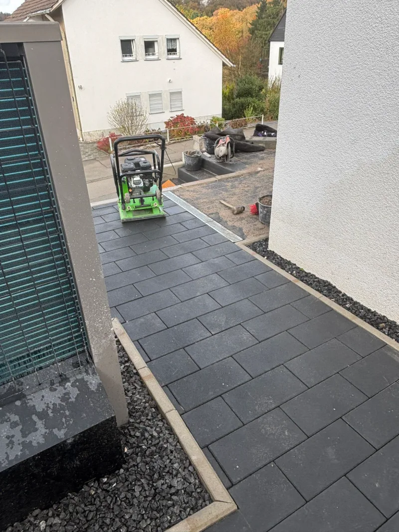 Frisch verlegter Weg aus dunklen Platten zwischen zwei Hauswänden, hinten eine grüne Rüttelplatte