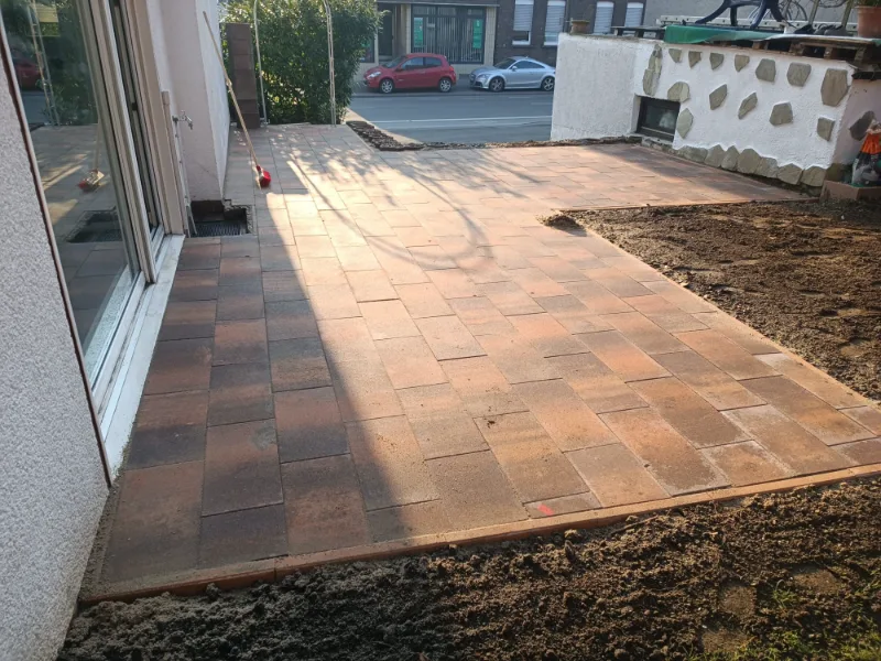 Terrasse aus rotbraunen Platten vor einer Fensterfront, daneben frisch vorbereitete Erde