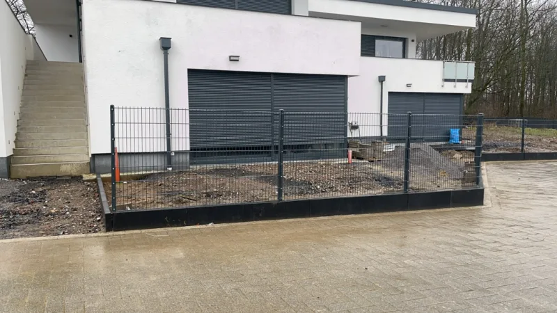Neubau mit Garagen, davor ein dunkler Stabmattenzaun auf einer Einfassung und ein Kiesstreifen, vorn die gepflasterte Zufahrt