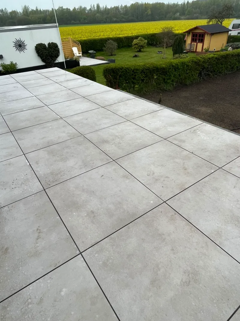 Terrasse aus hellen Großformatplatten, dahinter eine geschnittene Hecke, ein Gartenhaus und ein blühendes Feld