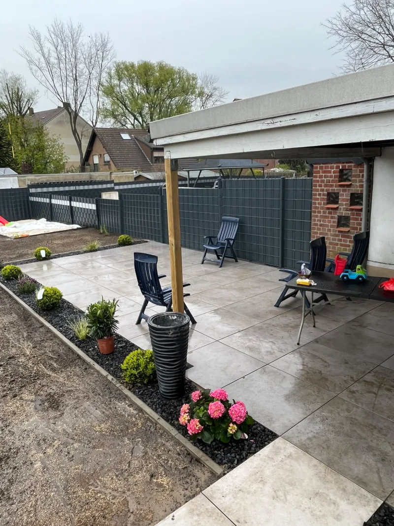 Terrasse aus hellen Platten unter einer Holzpergola, davor Gartenstühle, ein Kiesbeet mit Hortensien und ein Sichtschutzzaun