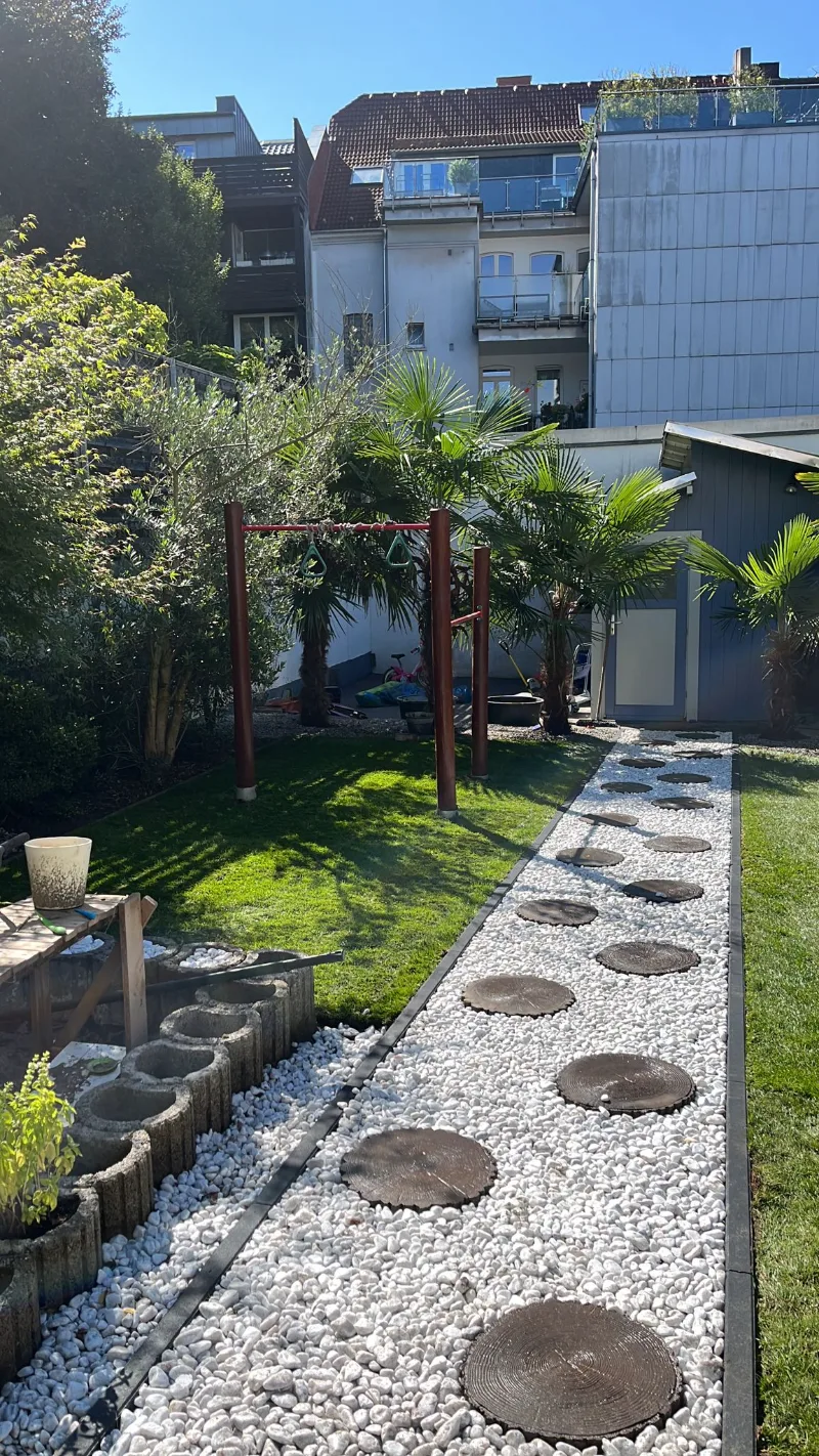 Runde Trittplatten in einem Streifen aus weißem Kies, daneben Rasen, Palmen und eine Pergola